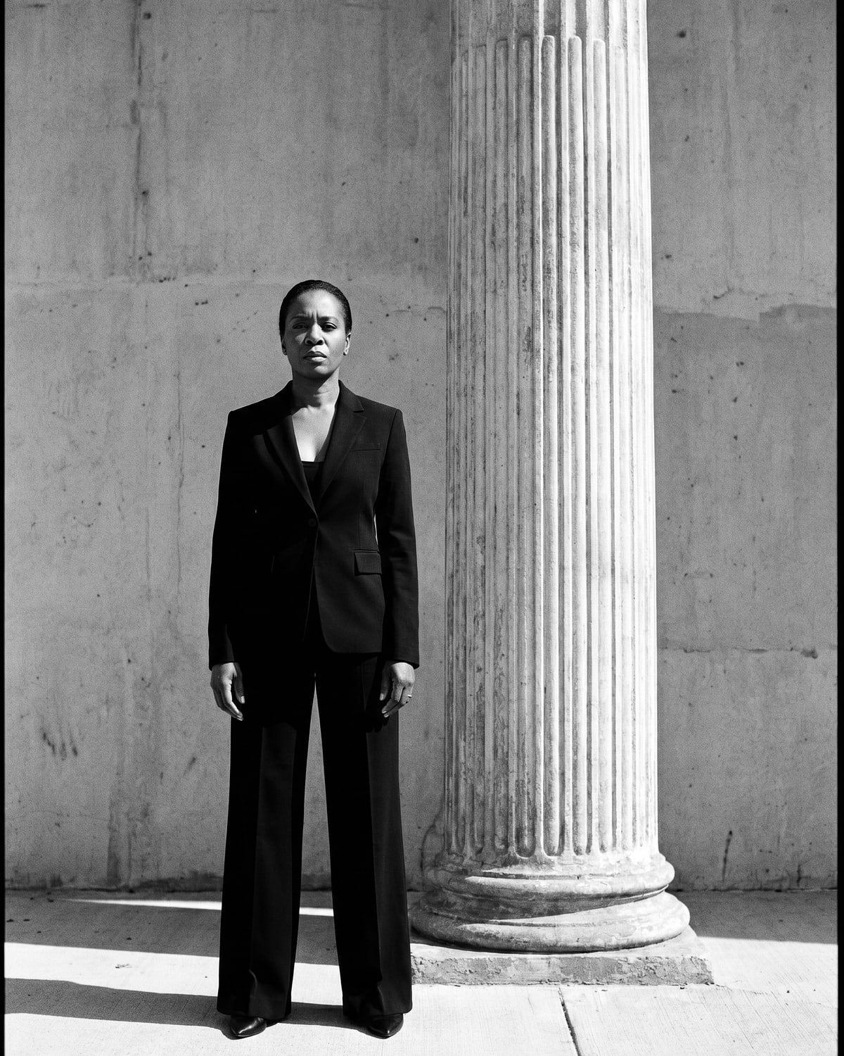 Black-and-white portrait of a woman in a structured blazer and trousers beside a classical fluted column.