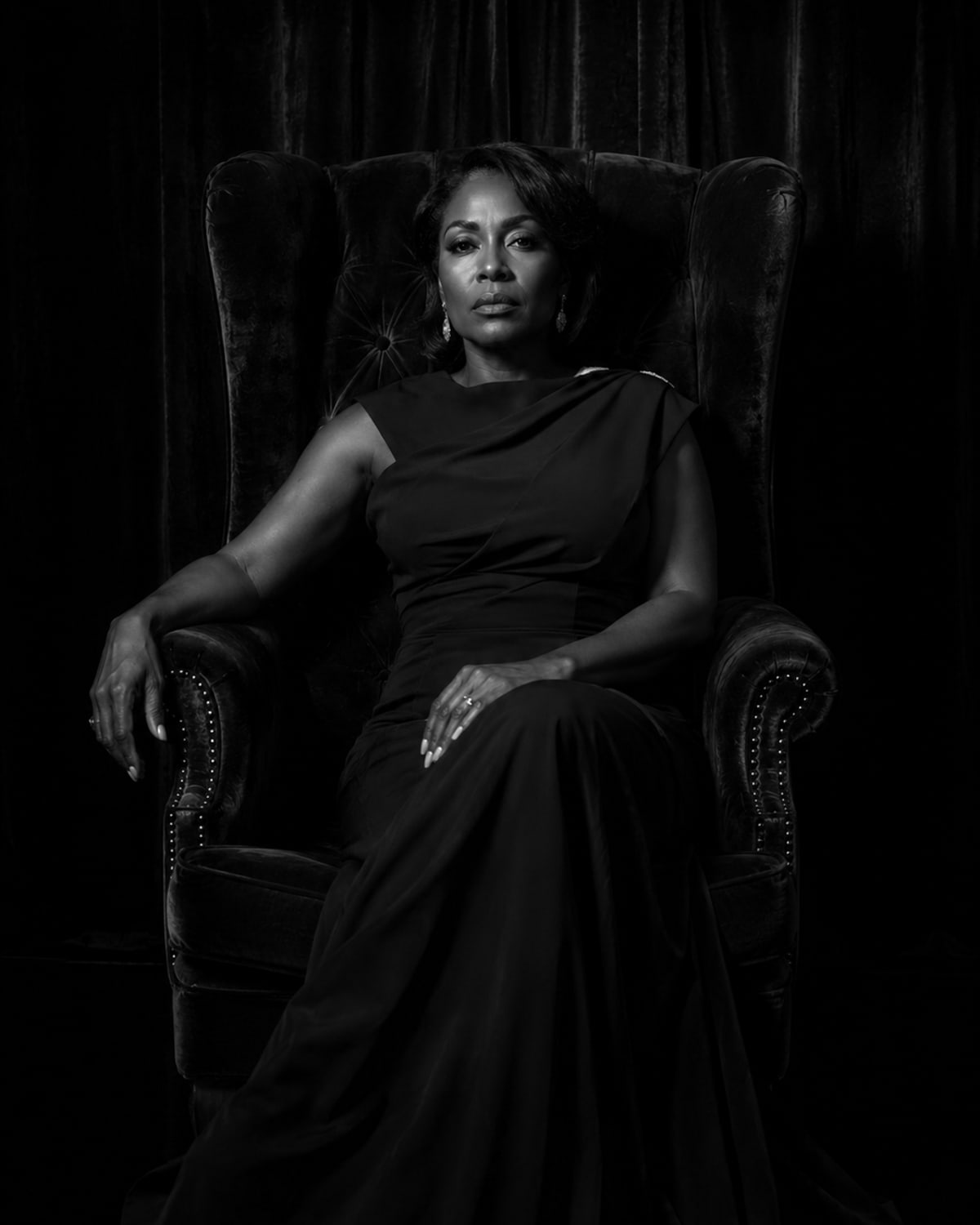 Black-and-white portrait of a woman in a floor-length black gown beside a tufted velvet wingback chair.